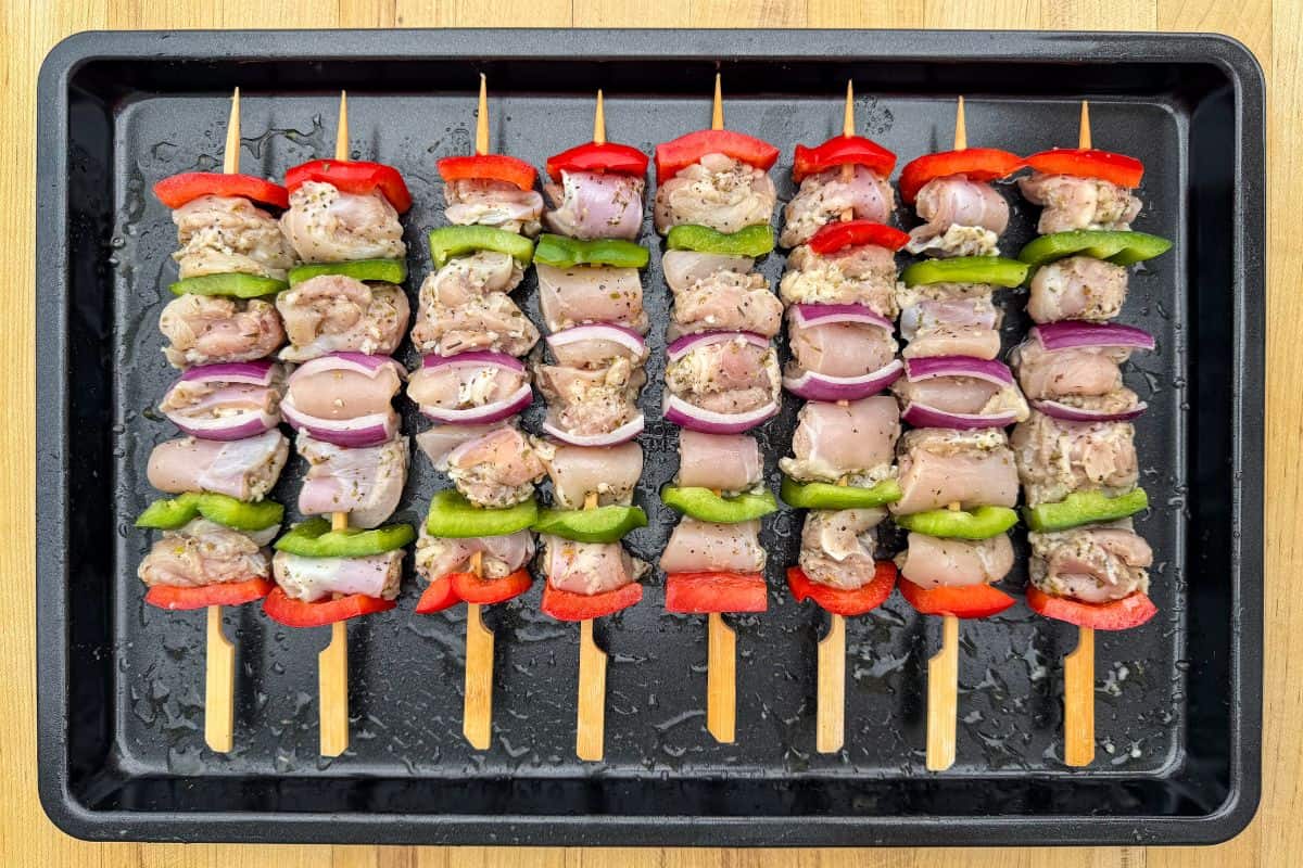 A sheet pan of chicken souvlaki skewers with vegetables are ready to get grilled.