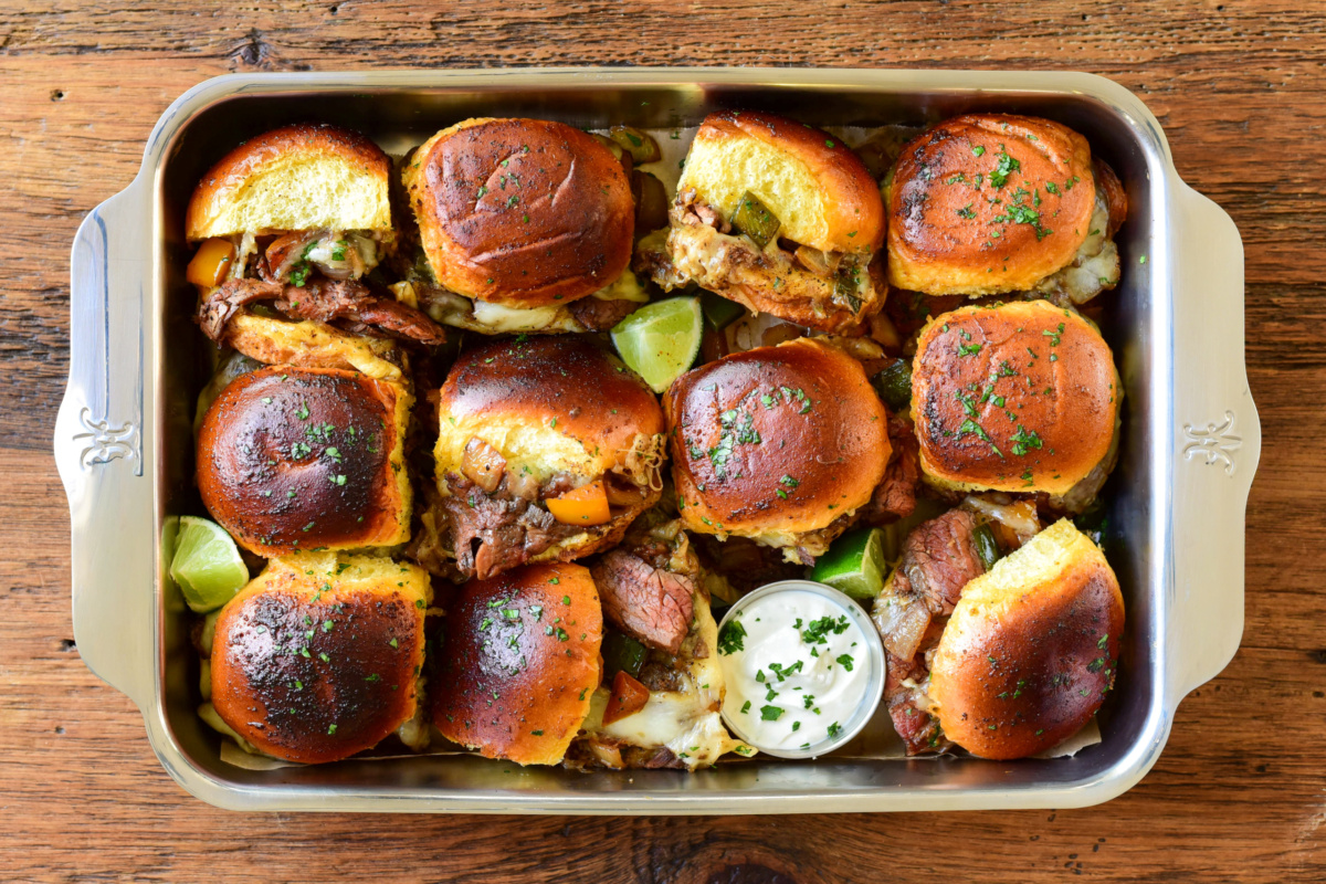 A pan with a dozenx steak sliders. Some of the sliders are on their sides, revealing the insides. They are served with a lime-crema and lime wedges.