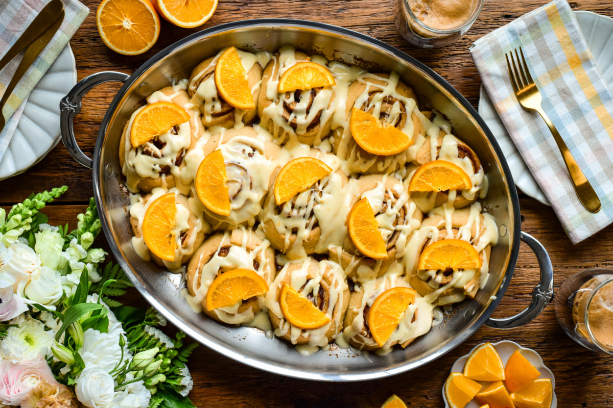 An oval au gratin roaster with orange glazed cinnamon rolls, each topped with a slice of candied orange. Orange slices, coffee and a floral bouquet surround the pan.