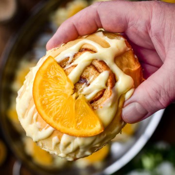 A close-up of a cinnamon roll with orange cream cheese glaze and a piece of candied orange.
