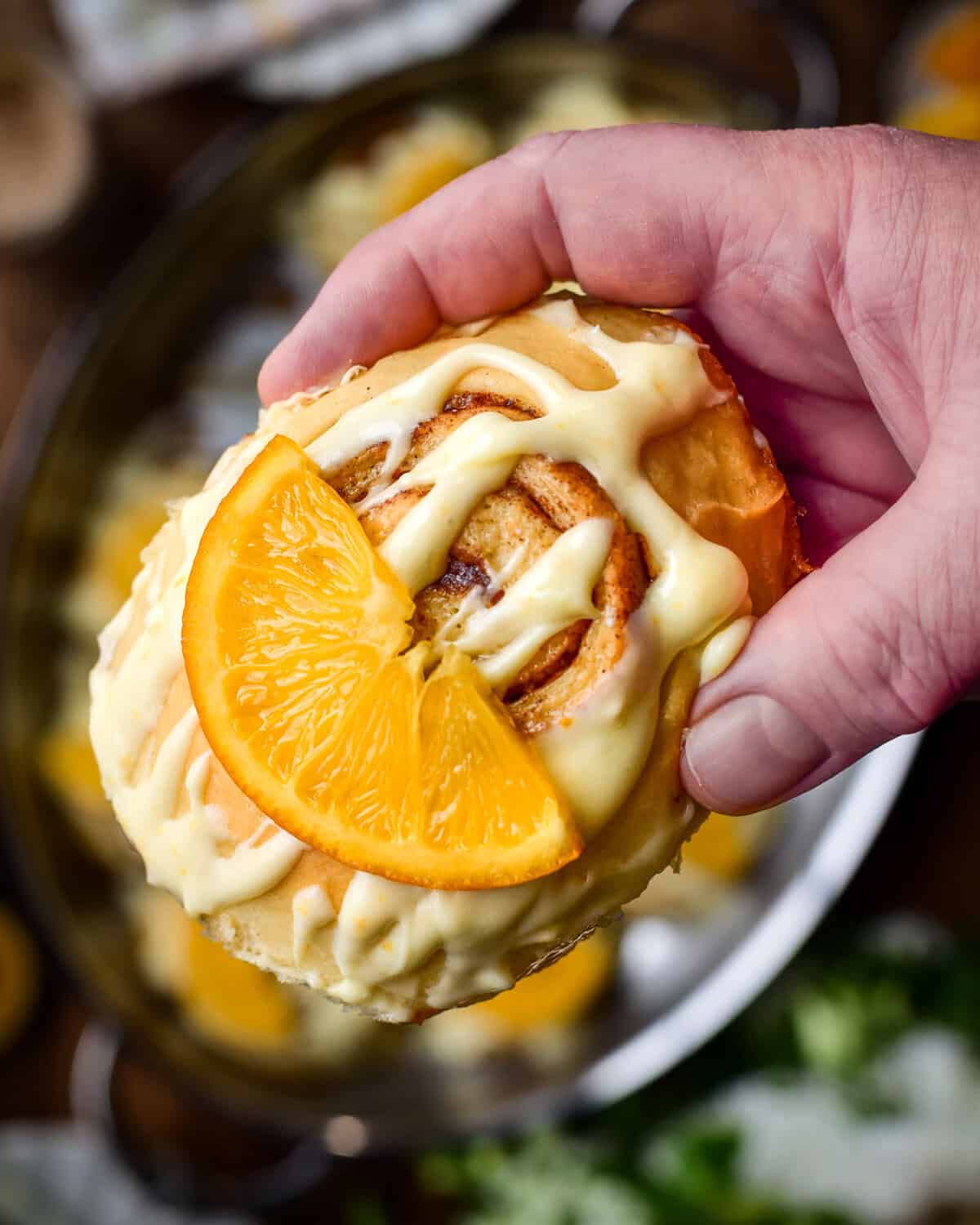 A close-up of a cinnamon roll with orange cream cheese glaze and a piece of candied orange.