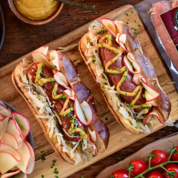 Two charcoal grilled bratwurst with pickled apples, sauerkraut, and mustard on pretzel buns.