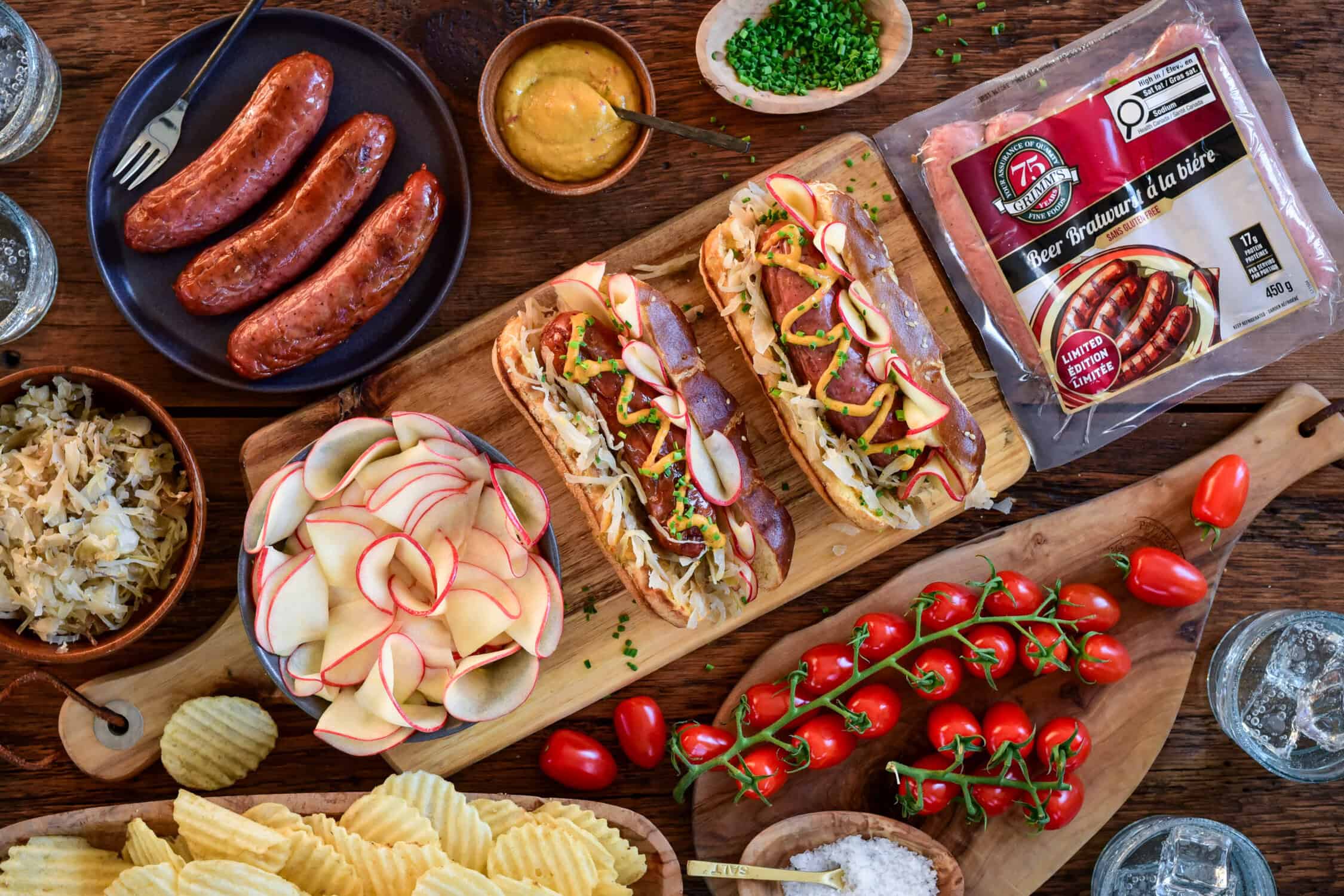 Two charcoal-grilled bratwurst with pickled apples, sauerkraut, and mustard on pretzel buns. Surrounded by extra ingredients to build more brats on pretzel buns.