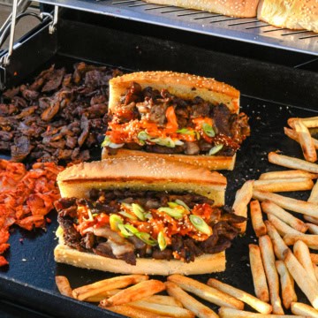 Two bulgogi cheesesteaks on the griddle with fries and extra sandwich ingredients.