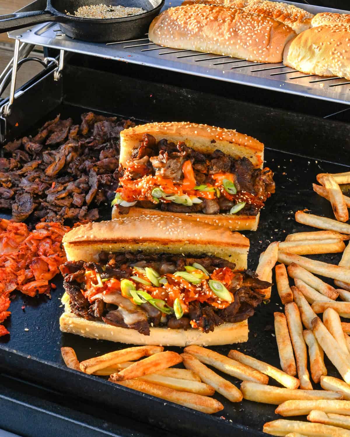 Two bulgogi cheesesteaks on the griddle with fries and extra sandwich ingredients.