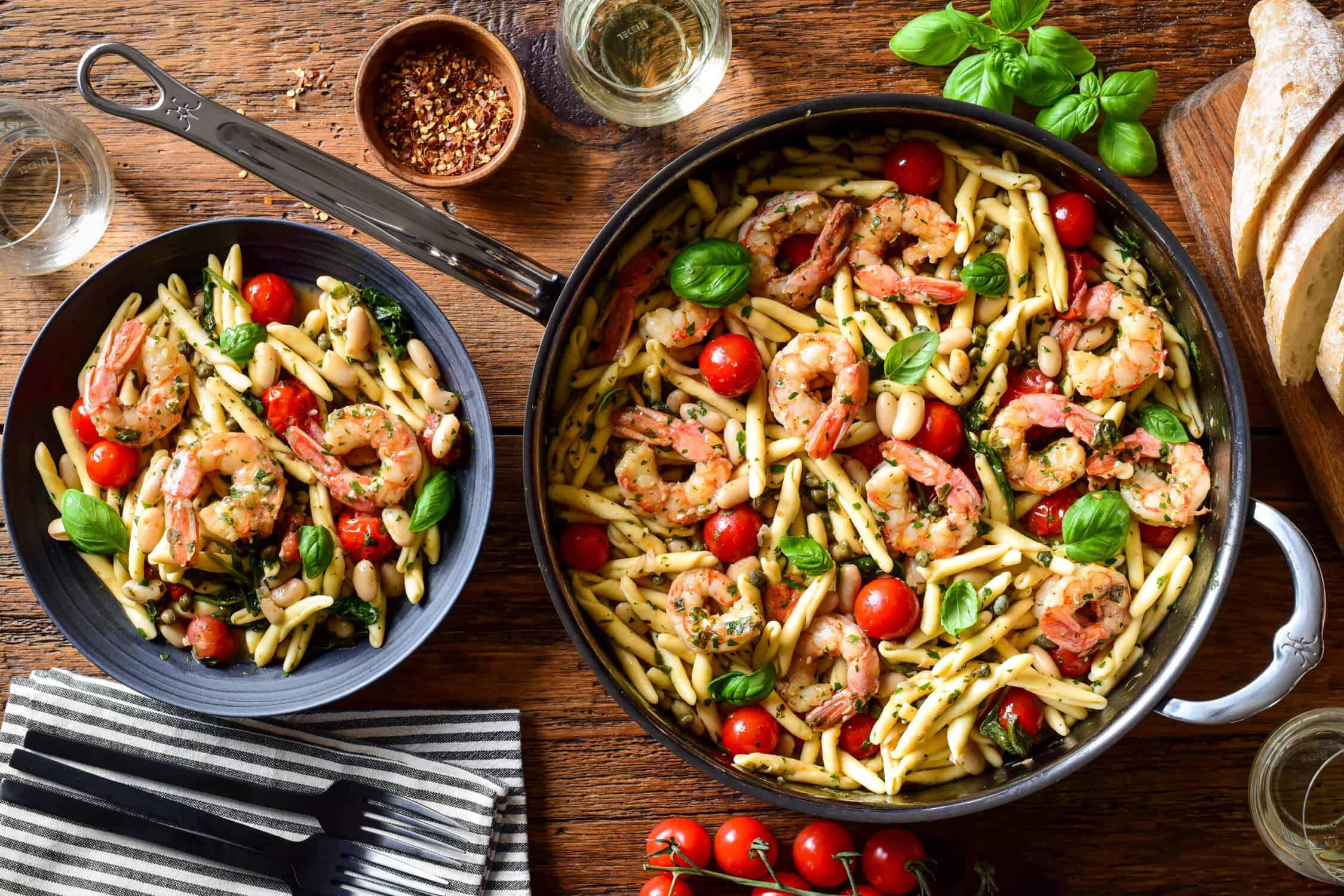 A pan and a bowl with Lemon Caper Shrimp Piccata Pasta with White Beans. This pasta is served with bread, chili flakes white wine an extra fresh basil and tomatoes.