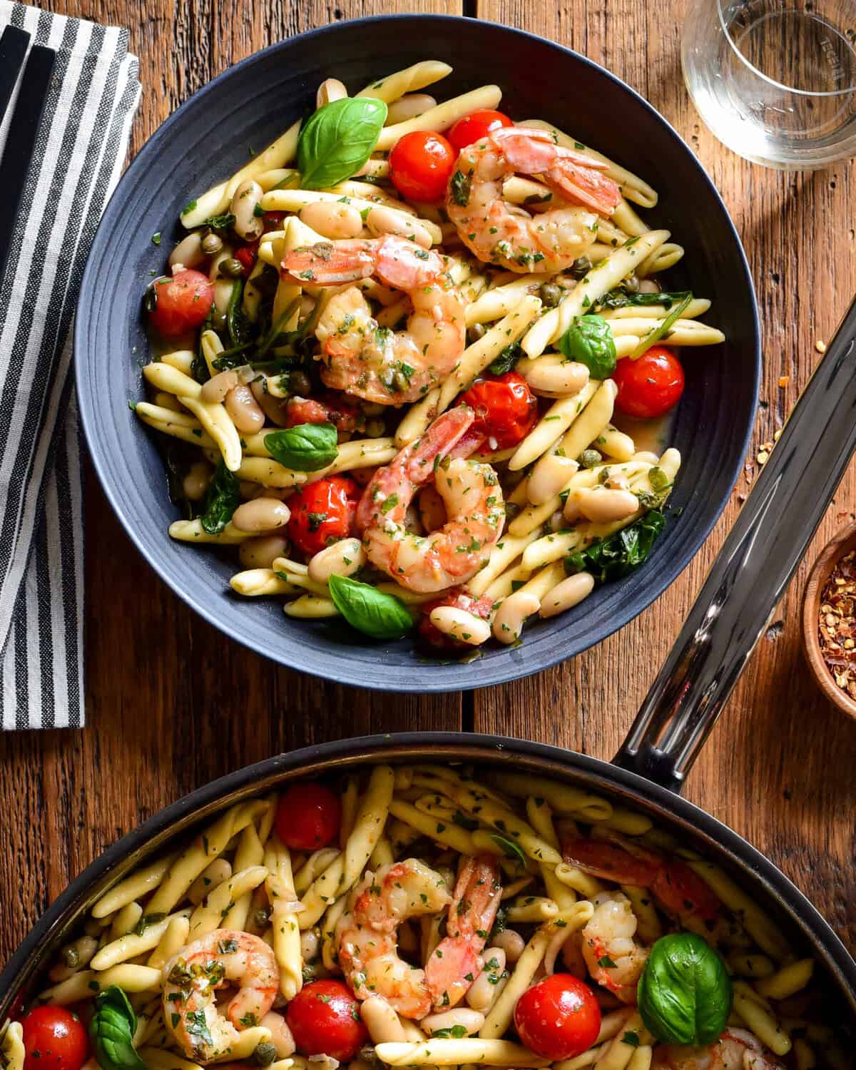 A pan and a bowl with Lemon Caper Shrimp Piccata Pasta with White Beans. This pasta is served with bread, chili flakes white wine an extra fresh basil and tomatoes.