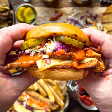 Close-up of a Nashville hot honey veal cutlet sandwich with pickles, coleslaw, and sauce in a bun, held by hands over a rustic wooden table with sides and drinks.