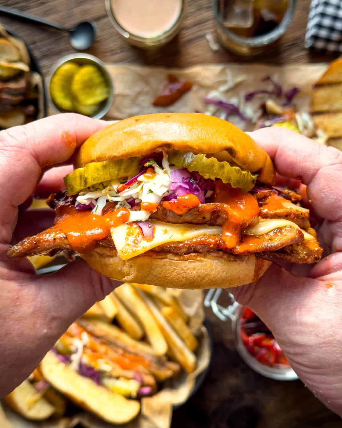 Close-up of a Nashville hot honey veal cutlet sandwich with pickles, coleslaw, and sauce in a bun, held by hands over a rustic wooden table with sides and drinks.
