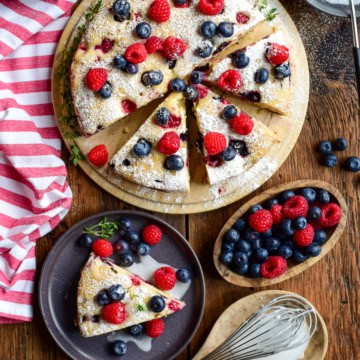A Lemon Blueberry and Raspberry Cake with blueberries and lemon drizzle on a round serving plate on a rustic wooden table with a red and white striped tea towel. A slice is served with extra berries and glaze, with extra berries on the side.