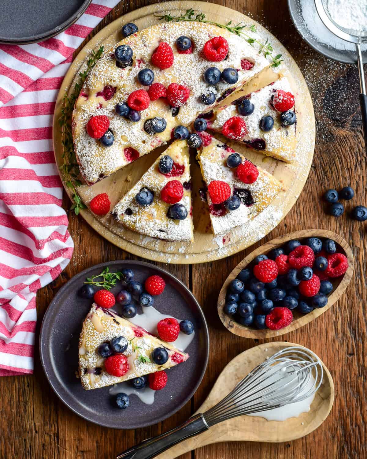A lemon raspberry cake with blueberries and lemon drizzle on a round serving plate on a rustic wooden table. A slice is served with extra berries and glaze, with extra berries on the side.