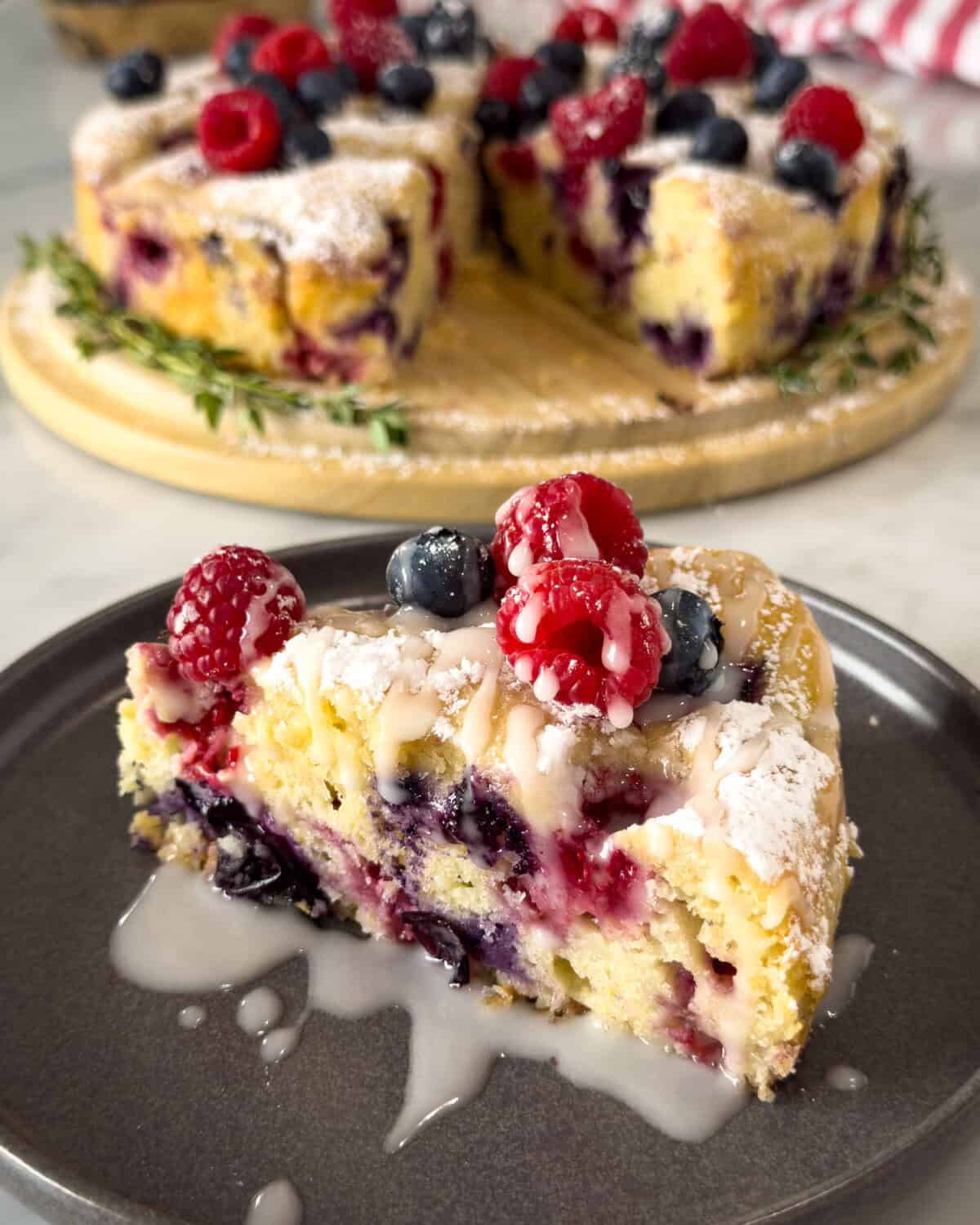 A slice of lemon, blueberry and raspberry cake topped with icing sugar and a lemon glaze. The cake is in the background.