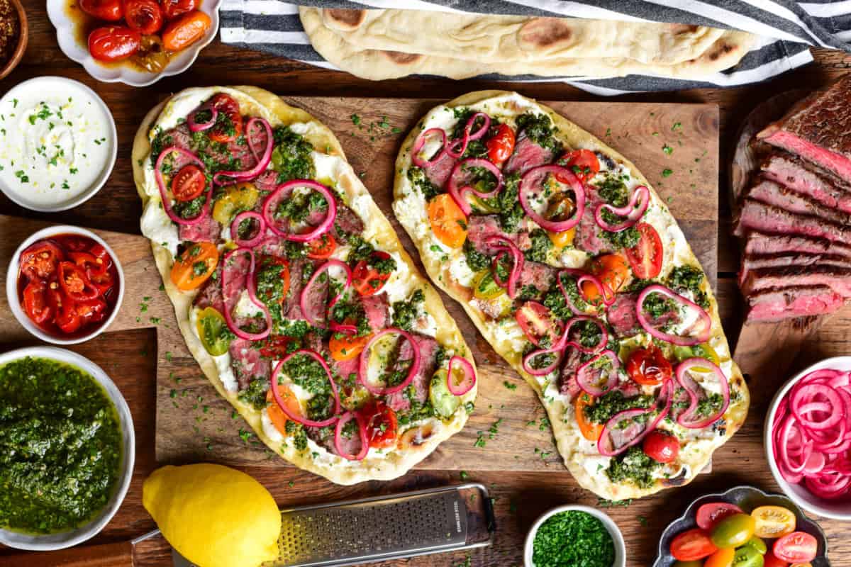 Two chimichurri steak flatbreads with whipped feta, marinated onions and blistered tomatoes on a wooden board. Extra ingredients are placed around this board.