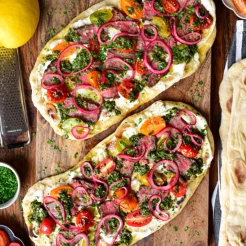 Two chimichurri steak flatbreads with whipped feta, marinated onions and blistered tomatoes on a wooden board. Extra ingredients are placed around this board.