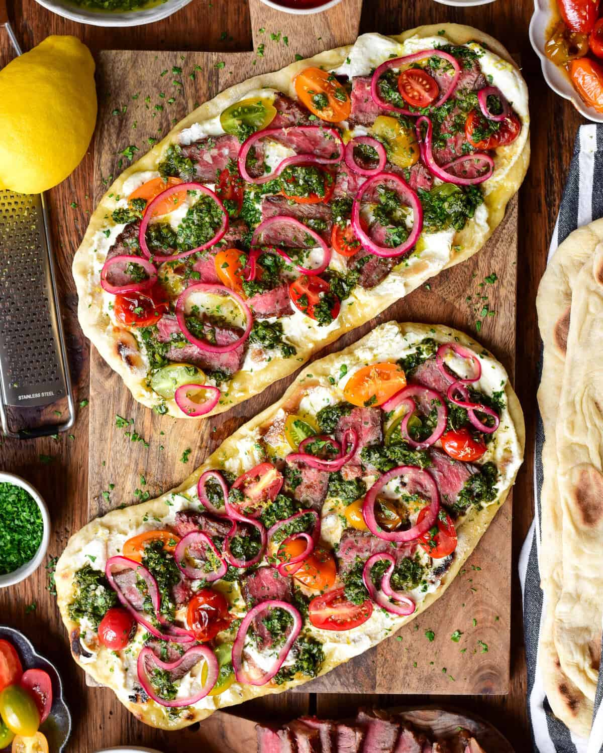 Two chimichurri steak flatbreads with whipped feta, marinated onions and blistered tomatoes on a wooden board. Extra ingredients are placed around this board.