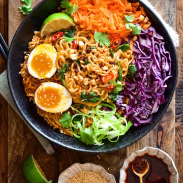 A colourful bowl of saucy chili-crunch almond butter ramen noodles in a large bowl with red cabbage, grated carrots and soft-boiled eggs. Extra chili crunch, lime wedges and toasted sesame seeds are served on the side.