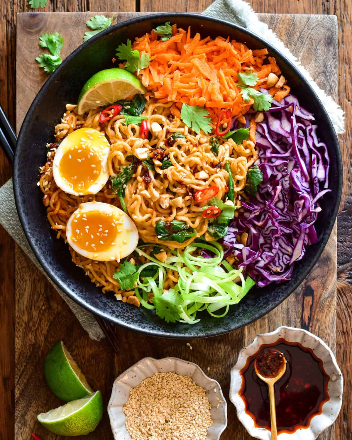 A colourful bowl of saucy chili-crunch almond butter ramen noodles in a large bowl with red cabbage, grated carrots and soft-boiled eggs. Extra chili crunch, lime wedges and toasted sesame seeds are served on the side.