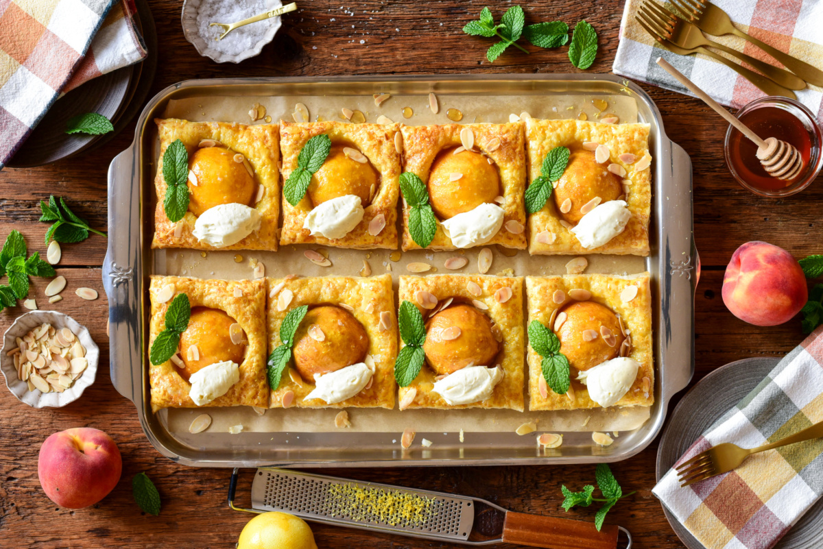 A tray with eight peach puff pastry tarts served with mint leaves and vanilla-almond mascarpone. Surrounded with extra mint leaves, fresh peaches, hot honey and finishing salt.