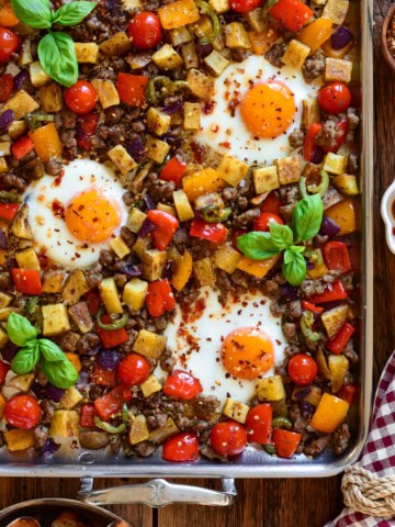 A stainless steel griddle with an Italian sausage and pepper breakfast hash with three fried eggs.