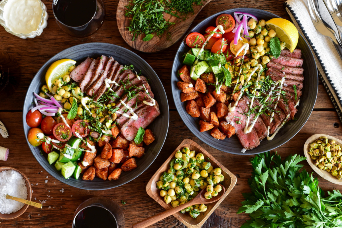 Two Mediterranean steak bowls with roasted sweet potato cubes, a chickpea salad, fresh tomatoes, fresh cucumbers, a lemon wedge, chopped herbs, a drizzle of a honey lemon tahini yogurt dressing and a lemon wedge.