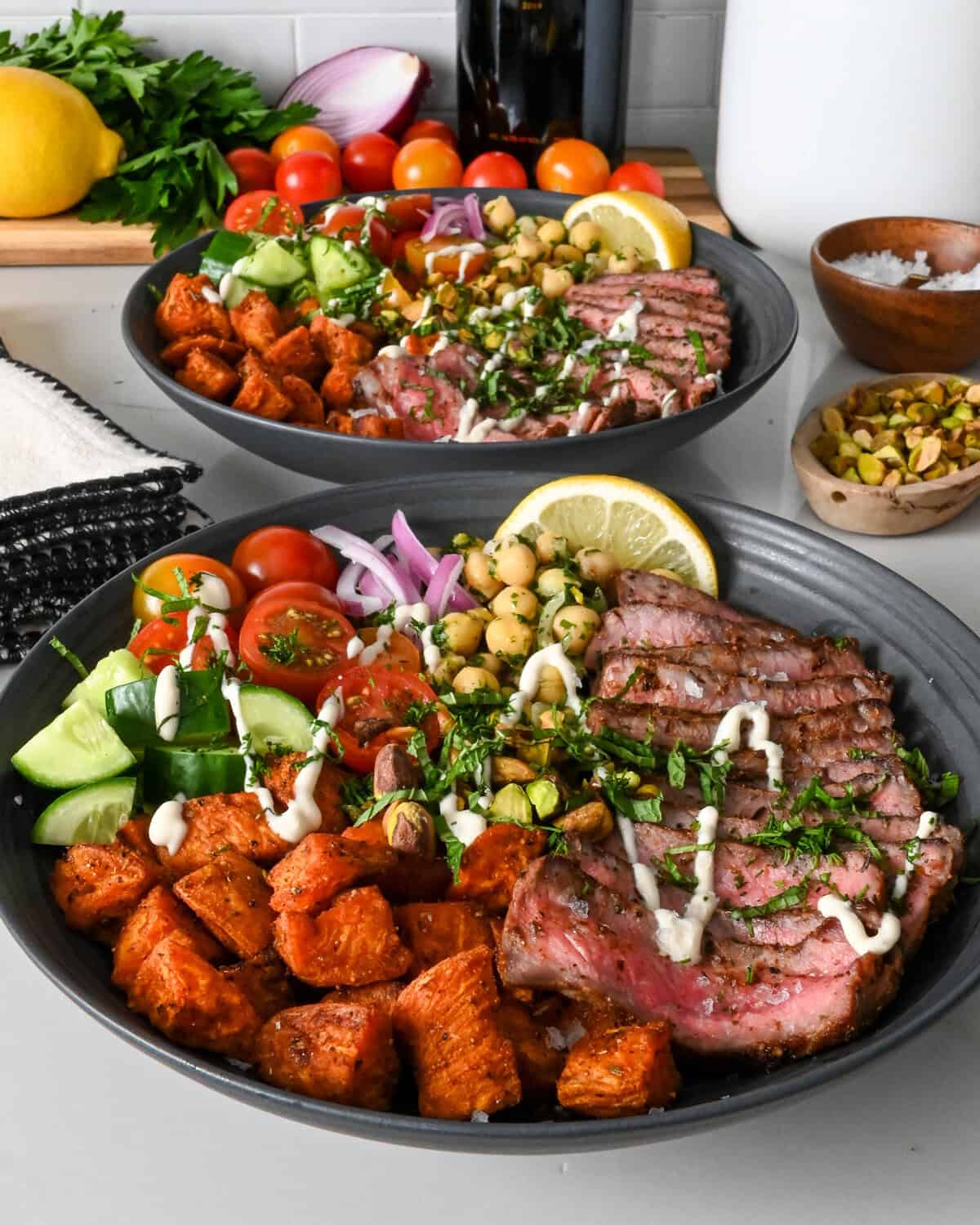 Two Mediterranean steak bowls with roasted sweet potato cubes, a chickpea salad, fresh tomatoes, fresh cucumbers, a lemon wedge, chopped herbs, a drizzle of a honey lemon tahini yogurt dressing and a lemon wedge.