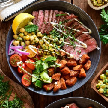 A Mediterranean steak bowl with roasted sweet potato cubes, a chickpea salad, fresh tomatoes, fresh cucumbers, a lemon wedge, chopped herbs, a drizzle of a honey lemon tahini yogurt dressing and a lemon wedge.