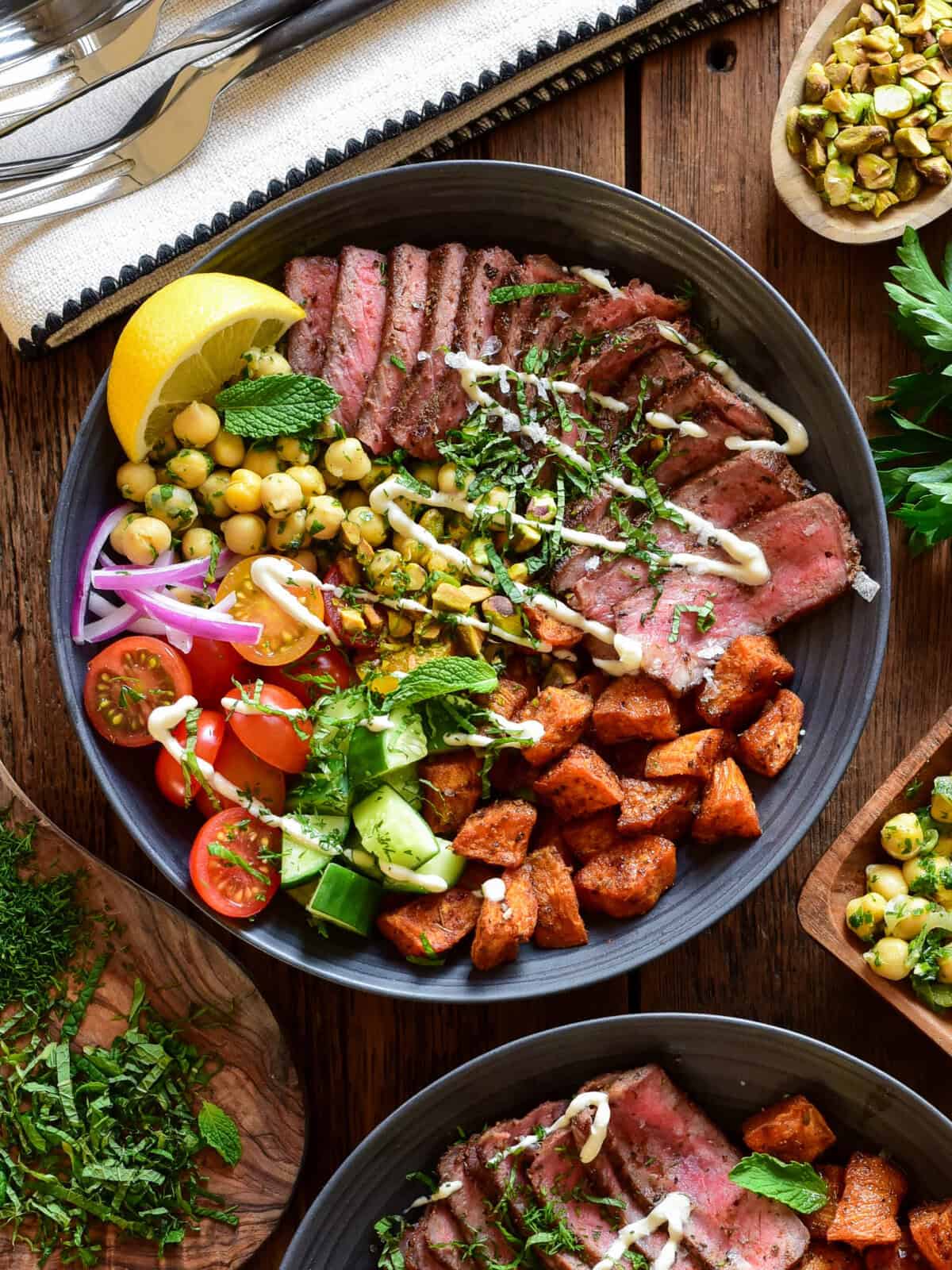 A Mediterranean steak bowl with roasted sweet potato cubes, a chickpea salad, fresh tomatoes, fresh cucumbers, a lemon wedge, chopped herbs, a drizzle of a honey lemon tahini yogurt dressing and a lemon wedge.