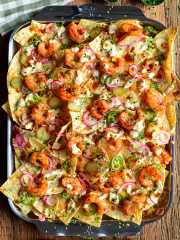 A tray of Nashville Hot Honey Shrimp Nachos with Pickle Crema. Layered with pickled onions, jalapenos, dill pickle slices and herbs.