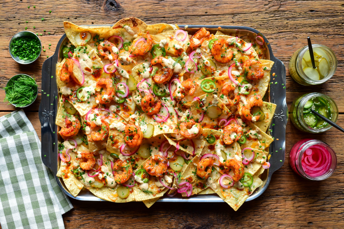 A tray of Nashville Hot Honey Shrimp Nachos with Pickle Crema. Layered with pickled onions, jalapenos, dill pickle slices, herbs and extra toppings on the side.