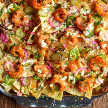 A close-up of a tray with Nashville Hot Honey Shrimp Nachos with Pickle Crema. Layered with pickled onions, jalapeños, dill pickle slices and herbs. Served with extra pickles on the side.