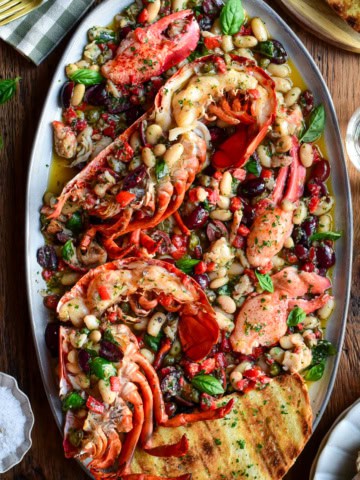 A colourful platter with charcoal-grilled lobster halves over warm Mediterranean garlic oil with beans, capers, olives, red peppers and herbs. It is served with toasted sourdough slices.