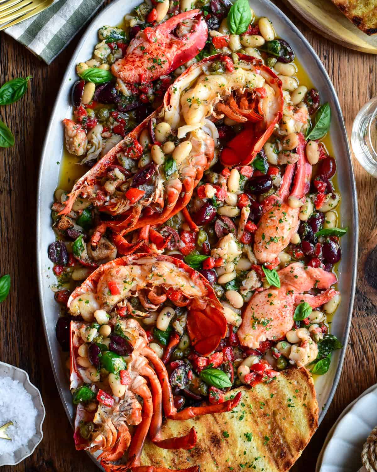 A colourful platter with charcoal-grilled lobster halves over a warm Mediterranean garlic oil with beans, capers, olives, red peppers and herbs. It is served with toasted sourdough slices.