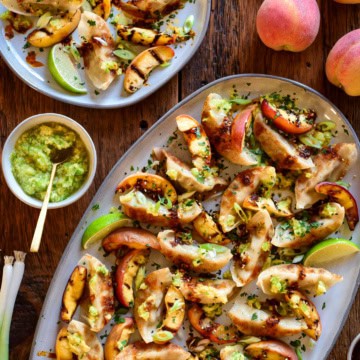 A crispy potsticker appetizer with charred peaches on a large oval platter. A portion is served with a lime wedge and extra scallion oil and fresh peaches are in this image.
