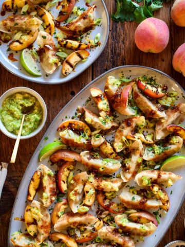 A crispy potsticker appetizer with charred peaches on a large oval platter. A portion is served with a lime wedge and extra scallion oil and fresh peaches are in this image.