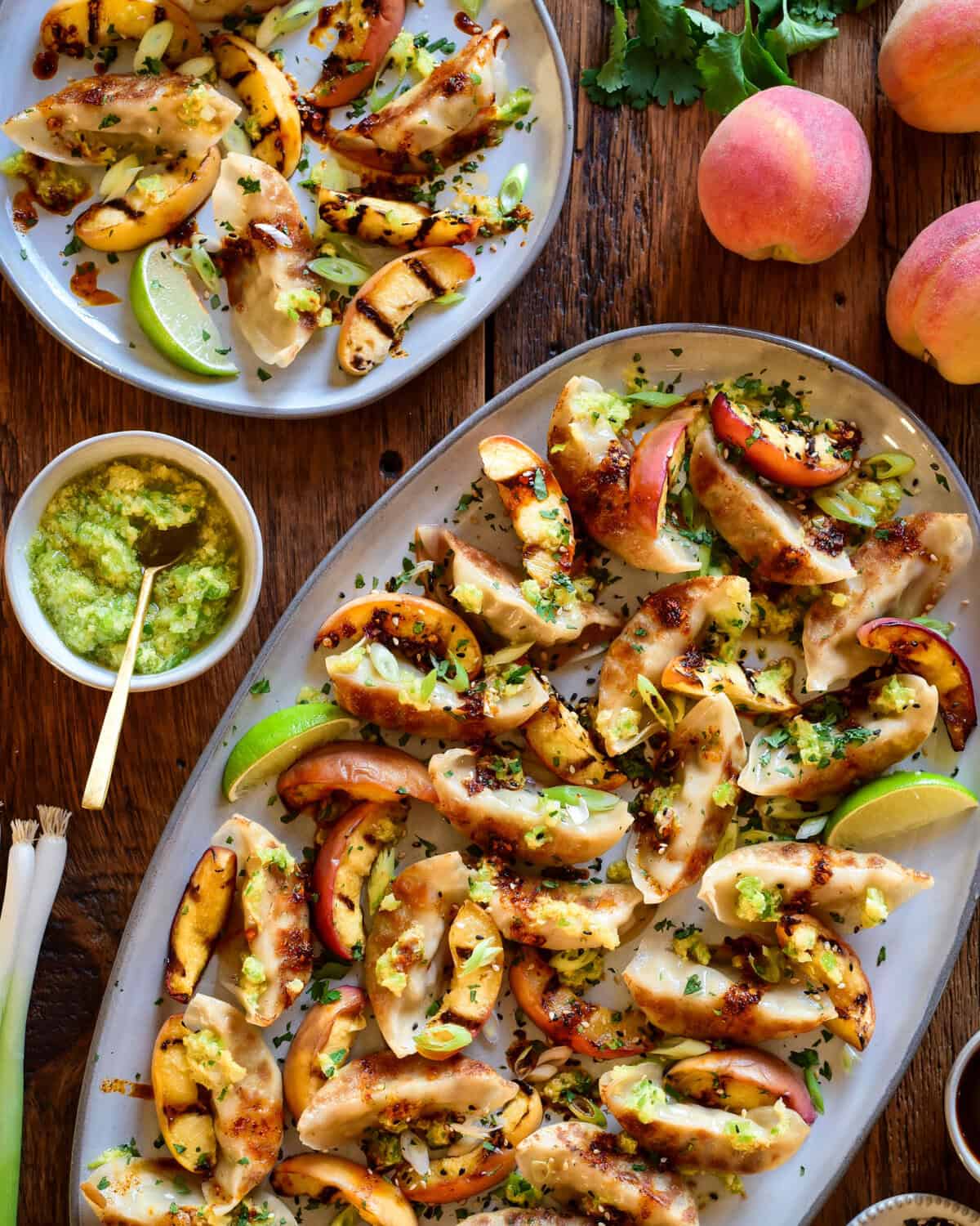 A crispy potsticker appetizer with charred peaches on a large oval platter. A portion is served with a lime wedge and extra scallion oil and fresh peaches are in this image.
