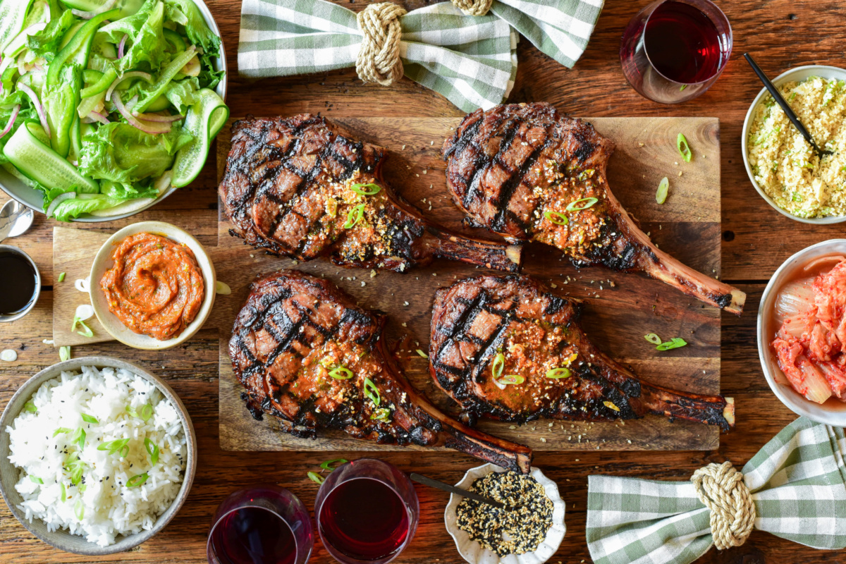 Four veal rib chops with distinct grill marks. They are placed on a wooden cutting board and served with extra gochujang butter, a Korean salad, kimchi and toasted sesame seeds. There are checkered napkins and a few red wine glasses.