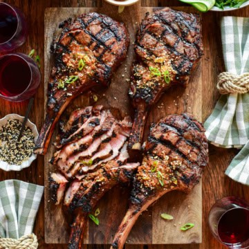 Four veal rib chops with distinct grill marks, one of which has been thinly sliced. They are placed on a wooden cutting board and served with extra gochujang butter, a Korean salad, kimchi and toasted sesame seeds. There are checkered napkins and a few red wine glasses.