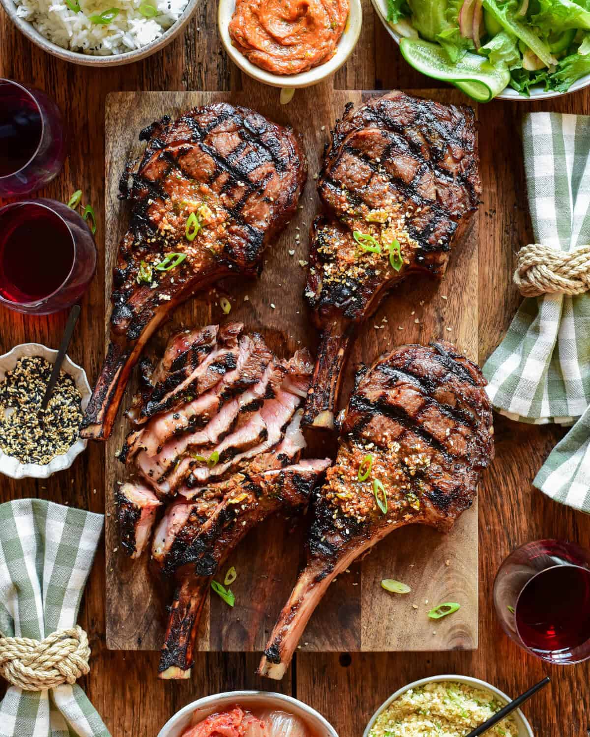 Four veal rib chops with distinct grill marks, one of which has been thinly sliced. They are placed on a wooden cutting board and served with extra gochujang butter, a Korean salad, kimchi and toasted sesame seeds. There are checkered napkins and a few red wine glasses.