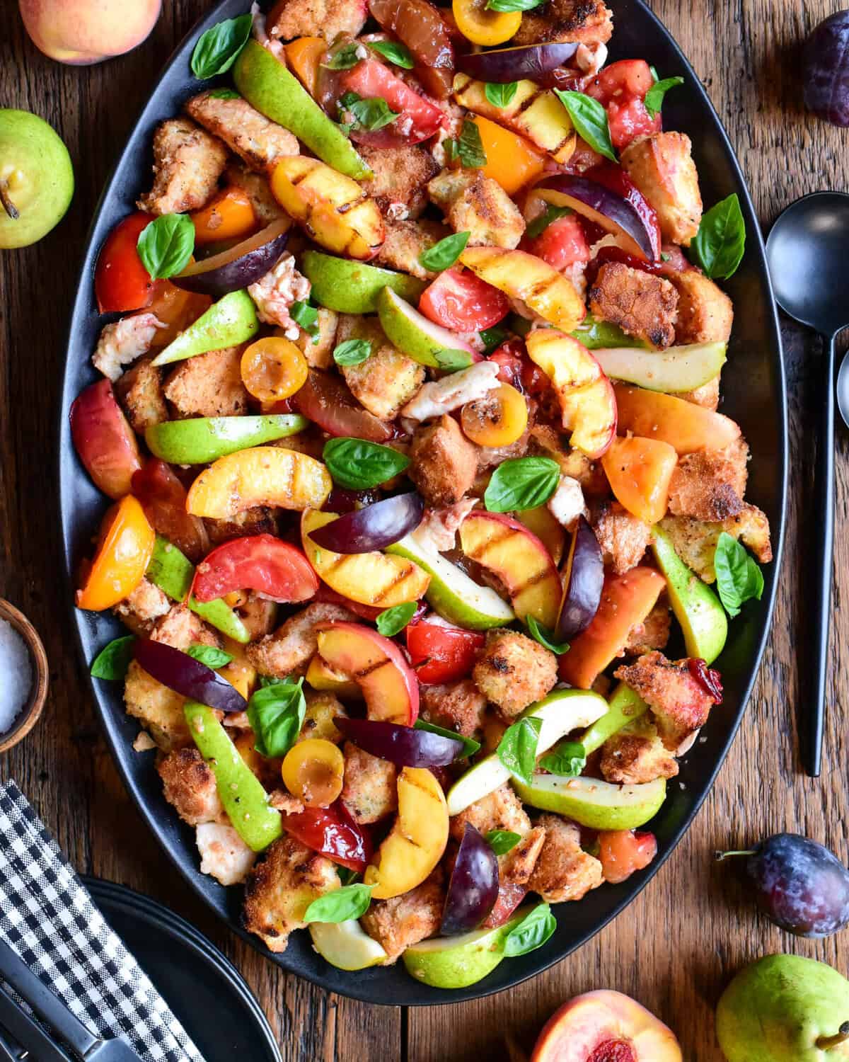 Colorful late summer stone fruit panzanella salad with fresh basil on a black platter, featuring peaches, plums, and tomatoes, perfect for seasonal dining.