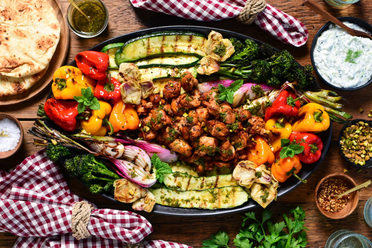 An oval platter with Mediterranean grilled chicken pieces in the center, surrounded by colourful grilled vegetables. Served with a whipped feta tzatziki, grilled flatbreads, drinks and checkered napkins.