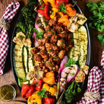 An oval platter with Mediterranean grilled chicken pieces in the center, surrounded by colourful grilled vegetables. Served with a whipped feta tzatziki, grilled flatbreads, drinks and checkered napkins.