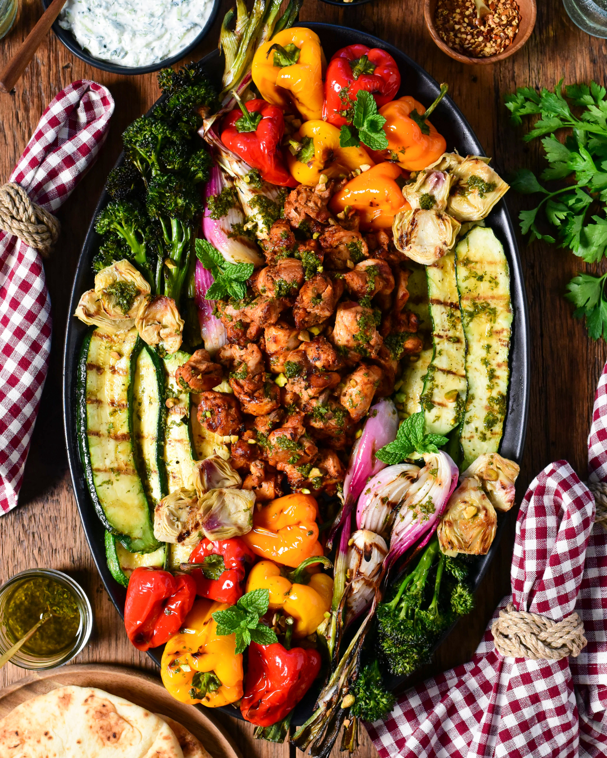 An oval platter with Mediterranean grilled chicken pieces in the center, surrounded by colourful grilled vegetables. Served with a whipped feta tzatziki, grilled flatbreads, drinks and checkered napkins.