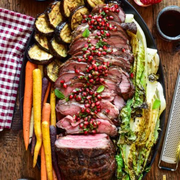 A top-down image of a smoked boneless leg of lamb that has been mostly sliced. A pomegranet mint relish is drizzled over the cut slices. Roasted karrots, eggplant and lettuce are surrounding the lamb on the plate.
