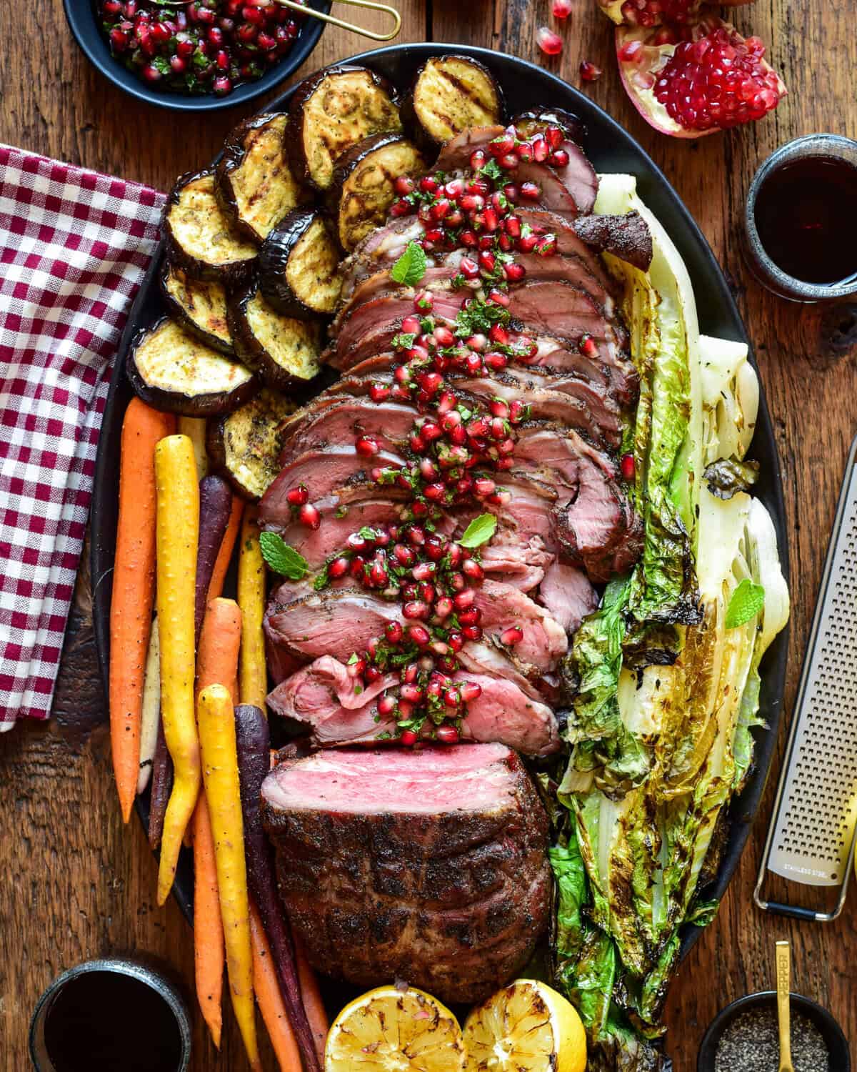 A top-down image of a smoked boneless leg of lamb that has been mostly sliced. A pomegranet mint relish is drizzled over the cut slices. Roasted karrots, eggplant and lettuce are surrounding the lamb on the plate.