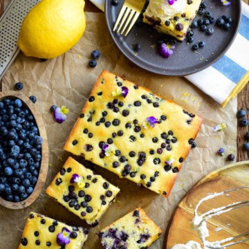 Freshly baked blueberry ricotta snack cake topped with whipped cream and edible flowers, served on a dark plate with blueberries and lemon nearby.