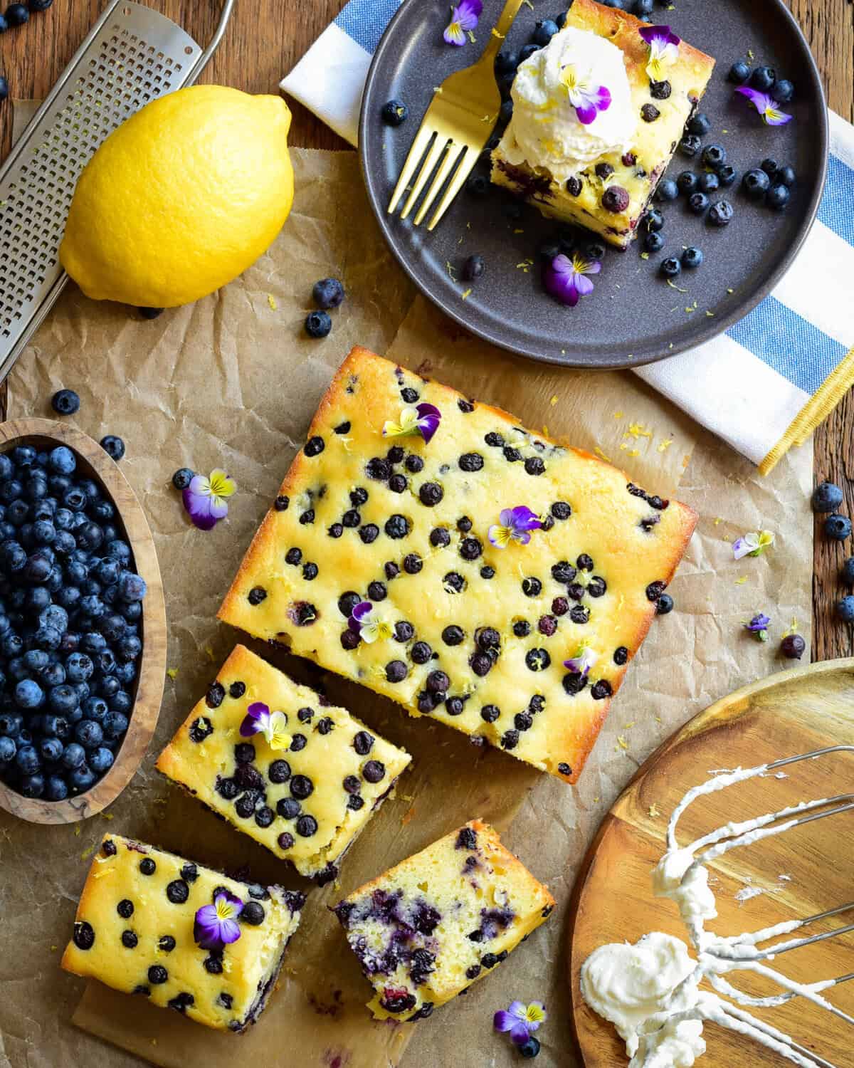 Freshly baked blueberry ricotta snack cake topped with whipped cream and edible flowers, served on a dark plate with blueberries and lemon nearby.
