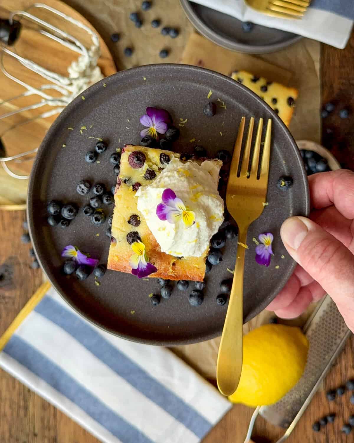 A top-down view of a slice of a wild blueberry ricotta cake topped with whipped cream, fresh blueberries and edible flowers.
