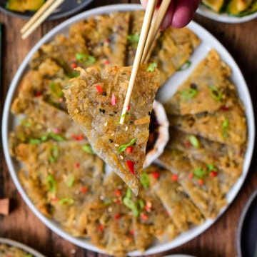 Close-up of crispy Korean Gamja-Jeon potato pancakes garnished with green onions and red pepper flakes, served on a white plate with dipping sauce.