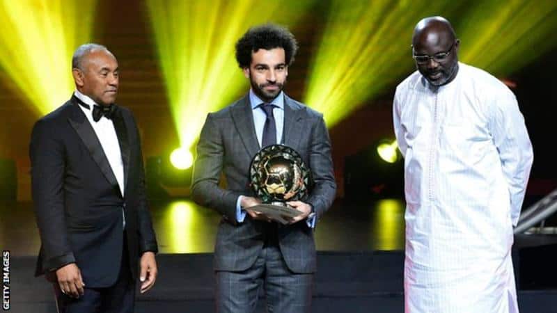 Award ceremony with three men on stage, one holding a trophy.