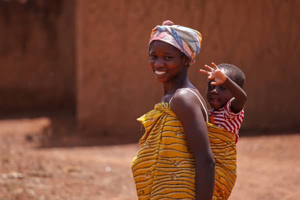 African woman with a child in traditional attire, smiling outdoors.